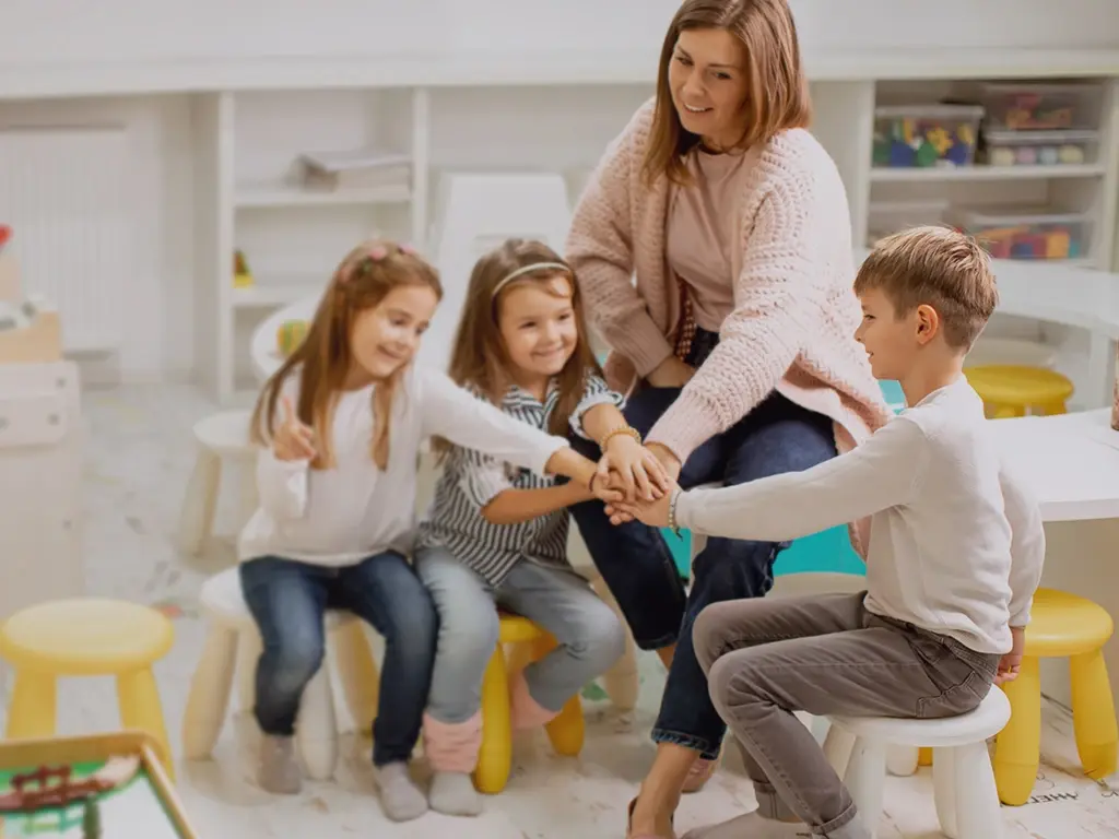 Eine Erzieherin mit drei Kindern, die ihre Hände aufeinander legen.